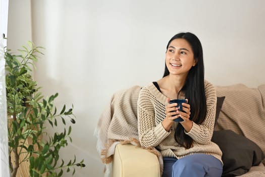 Calm young woman in sweater with a hot cup of coffee sitting on sofa in living room.