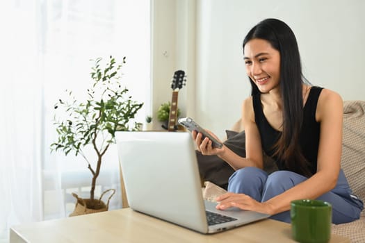 Cheerful young woman working from home with laptop and using mobile phone.