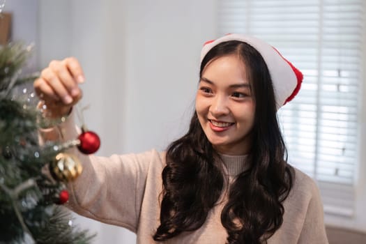 A young woman wearing a Santa hat decorates a Christmas tree with ornaments, capturing the festive spirit in a cozy home environment.