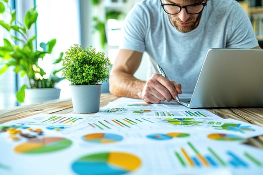 A focused businessman works diligently on financial charts and data in a stylish workspace, using a laptop and surrounded by greenery for inspiration and productivity.