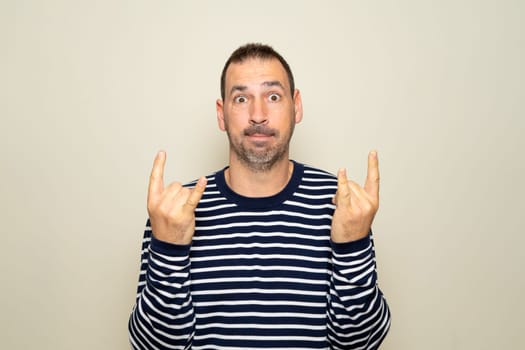 Portrait of bearded man showing rock and roll gesture, heavy metal sign, exclaiming with joy. Indoor studio photo isolated on beige background