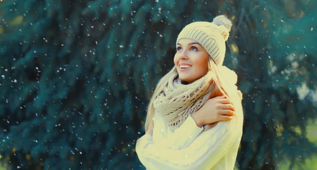 Winter portrait of happy smiling blonde young woman in forest with snow on Christmas tree background, wearing hat, enjoying warm weather, knitted sweater and scarf