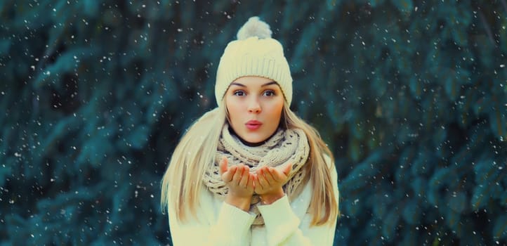 Happy young woman in winter park blowing kiss against Christmas tree background with snow, girl in white hat, in snowy forest