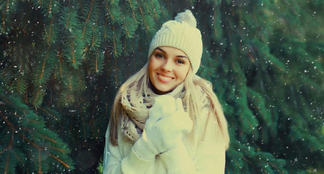 Winter portrait of happy smiling blonde young woman in forest with snow on Christmas tree background, wearing hat, enjoying warm weather, knitted sweater and scarf