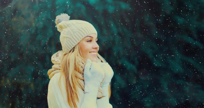 Winter portrait of happy smiling blonde young woman in forest with snow on Christmas tree background, wearing hat, enjoying warm weather, knitted sweater and scarf