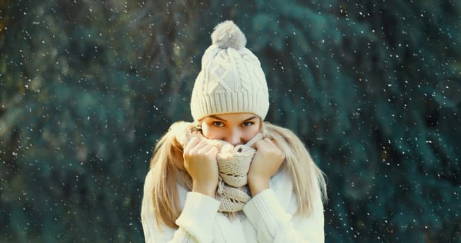 Winter portrait of happy smiling blonde young woman in forest with snow on Christmas tree background, wearing hat, enjoying warm weather, knitted sweater and scarf