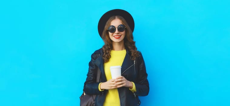 Portrait of stylish happy young woman holding cup of coffee in black round hat, leather jacket with backpack on bright blue background