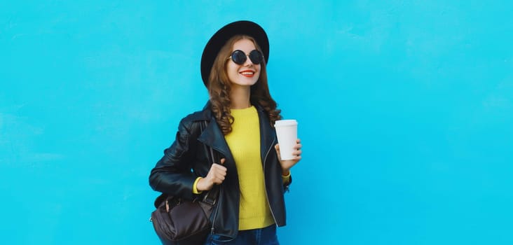 Portrait of stylish happy young woman holding cup of coffee in black round hat, leather jacket with backpack on bright blue background