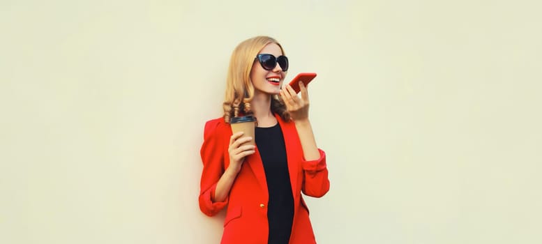 Beautiful young woman calling on smartphone in red business suit, holding cup of coffee standing on white background