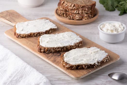 Slices of whole grain bread topped with cream cheese on a wooden cutting board in a cozy kitchen setting.
