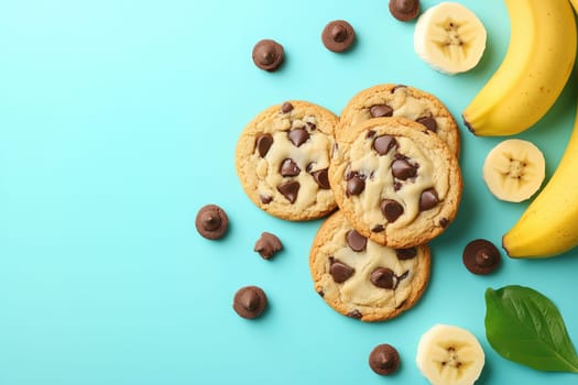 A delightful arrangement of freshly baked chocolate chip cookies sits next to ripe bananas and chocolate drops on a bright blue surface, showcasing a tasty snack.