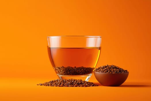 A clear glass filled with golden flaxseed tea sits next to a small bowl of whole flaxseeds. The backdrop features a bright orange color, enhancing the warmth of the beverage and seeds.