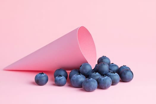 A paper cone in soft pink color is tipped over, spilling fresh blueberries onto a pastel surface. The bright display creates a vibrant and inviting atmosphere.