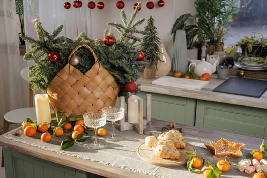 A cozy kitchen decorated for family holidays, with a wicker basket filled with fir branches and red ornaments. The table is decorated with tangerines, two elegant glasses and a variety of pastries