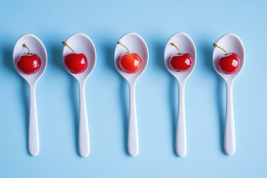 Five white porcelain spoons each hold a shiny red cherry, arranged neatly on a calming blue surface illuminated by natural light.