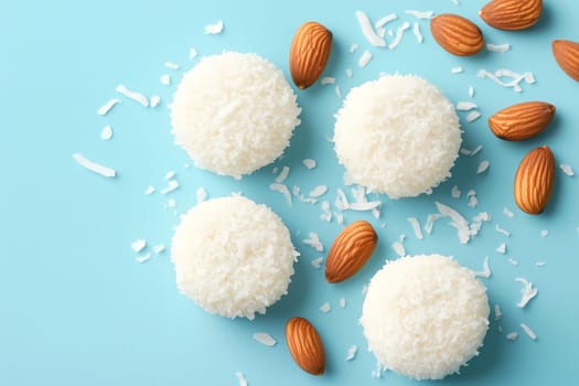 Four coconut balls are arranged beautifully on a light blue surface, surrounded by almonds and sprinkled coconut flakes, creating an enticing dessert display.