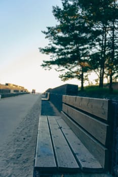 Close-up of an empty wooden bench facing a coastal pathway with trees and soft evening light. Tranquil outdoor setting with a peaceful atmosphere at sunset