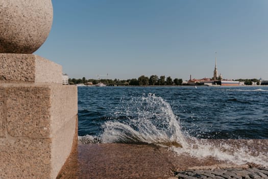 Close-up of waves crashing against a granite embankment on a sunny day, with a historic cityscape in the background under a clear blue sky. Vibrant waterfront scene
