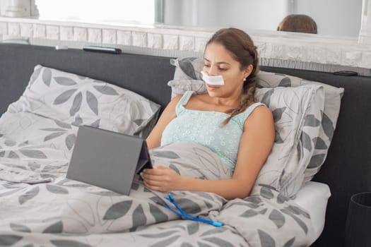 Woman resting in bed in casual clothes and bandages on her face after plastic surgery on her nose,using tablet to communicate with family, calm atmosphere and soft colors illustrate recovery process