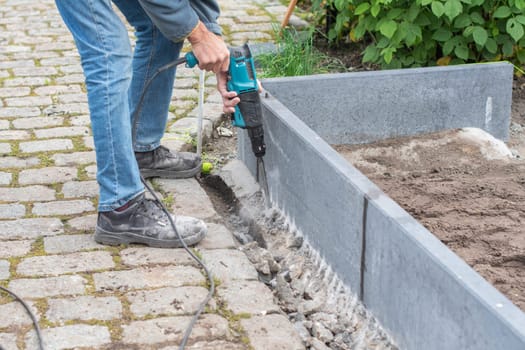middle-aged man drills holes in the foundation of greenhouse for strengthening, improvement of garden or vegetable garden,frame for beds with soil,concept of home ecological turning of vegetables,