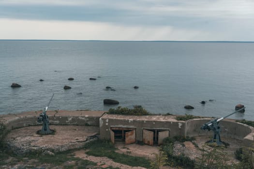 An abandoned coastal artillery bunker with cannons overlooks a calm sea under an overcast sky, evoking a sense of historical significance and quiet solitude in a rugged coastal landscape
