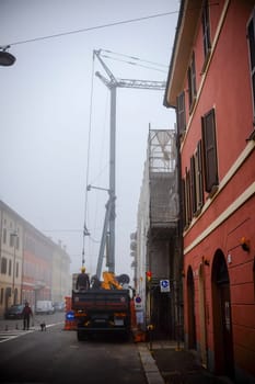 Cremona, Lombardy, Italy - November 7th 2024 Construction crane and truck parked on a city street on a foggy day, with construction workers and scaffolding visible
