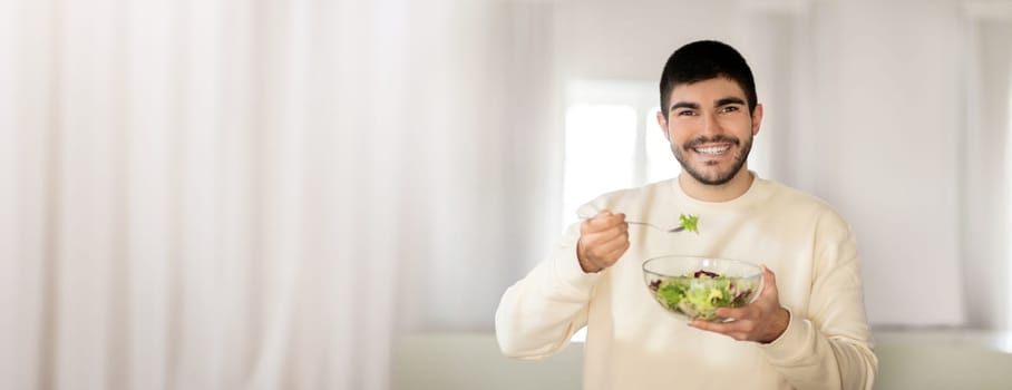 A smiling young man holds a bowl of fresh salad while taking a bite. The bright indoor space enhances the healthy lifestyle vibe, highlighting freshness and good nutrition.