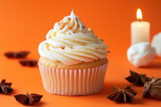 A beautifully decorated vanilla cupcake sits prominently on an orange surface, topped with rich creamy frosting and spices, alongside a flickering candle, creating a warm ambiance.