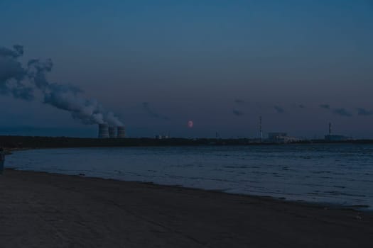 Industrial coastline at dusk with smoke stacks emitting smoke into the sky, viewed from a beach. Calm water and distant horizon create a moody, atmospheric scene