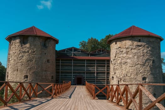 A grand entrance to an ancient fortress, featuring twin stone towers with red roofs connected by a wooden bridge under a clear blue sky. Iconic medieval architecture