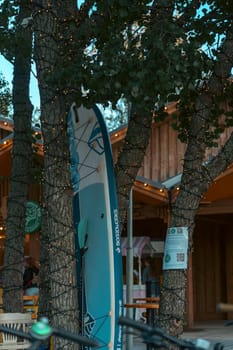 Blue surfboard propped against a tree near a rustic outdoor cafe, with warm lights creating a cozy atmosphere in the early evenin
