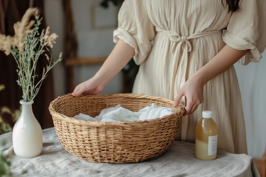 A woman in a flowing beige dress gently lifts a handcrafted wicker basket filled with freshly laundered clothes. The cozy environment features soft lighting, a vase of flowers, and a bottle nearby.