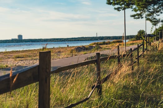 A coastal path bordered by a wooden fence runs alongside a grassy shoreline, offering a peaceful and scenic view of the ocean under a bright blue sky