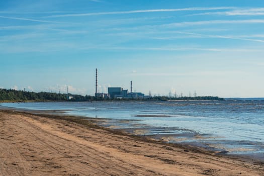A power plant stands along a sandy beach under a clear blue sky, juxtaposing industrial structures with the natural coastal landscape on a sunny day