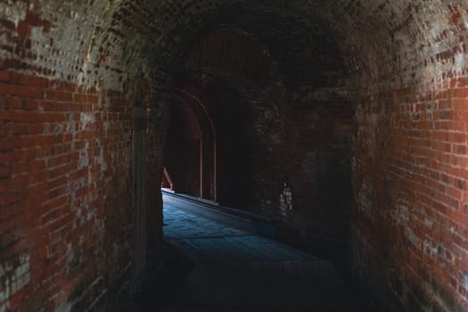 Interior of a dark, narrow brick tunnel with an arched ceiling inside a historic fortress. The dim lighting and rough texture create a mysterious, ancient atmosphere