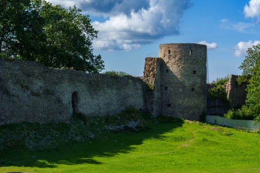 A medieval stone fortress with a tall round tower and extensive walls, set against a clear blue sky. The structure stands on a grassy area, evoking historical significance and rustic charm