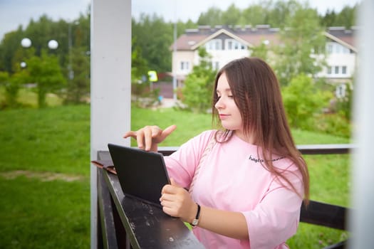A woman smiles as she looks at her tablet, sitting on porch with a grassy view and houses in the background