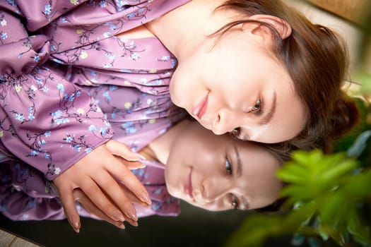 The woman reflects on her thoughts while resting her chin on her hand, surrounded by greenery in a cozy indoor setting.