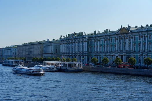 Elegant row of historic buildings along the river with boats docked by the waterfront. Clear blue sky and calm water create a tranquil urban riverside scene