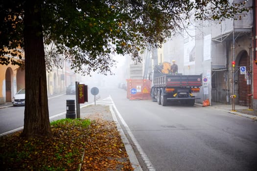 Cremona, Lombardy, Italy - November 7th 2024 Construction truck blocking street on a foggy autumn day, with a construction worker standing on the back