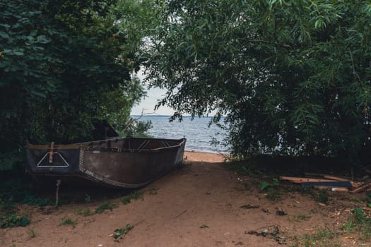 An old wooden boat rests on a sandy shore, partially hidden among trees, offering a secluded and peaceful setting by the water under an overcast sky, capturing a rustic lakeside charm