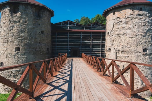 A grand entrance to an ancient fortress, featuring twin stone towers with red roofs connected by a wooden bridge under a clear blue sky. Iconic medieval architecture