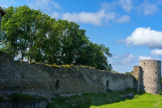 A medieval stone fortress with a tall round tower and extensive walls, set against a clear blue sky. The structure stands on a grassy area, evoking historical significance and rustic charm