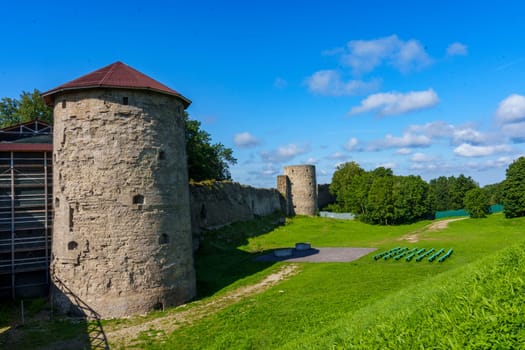 A medieval stone fortress with a tall round tower and extensive walls, set against a clear blue sky. The structure stands on a grassy area, evoking historical significance and rustic charm