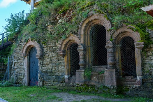 A close-up of an ancient stone facade featuring arched windows and overgrown with moss and vegetation, highlighting the weathered, historic architecture of a medieval fortress