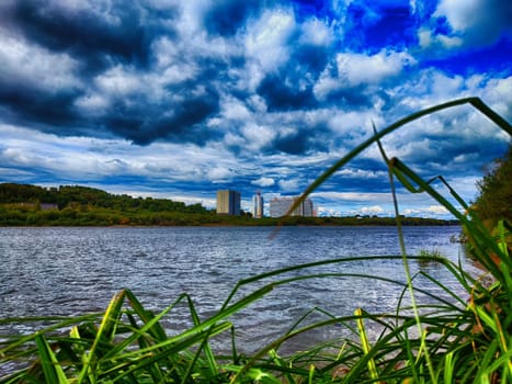 A tranquil afternoon shows a river bordered by grass, with urban buildings rising against a backdrop of a dramatic, cloudy sky.