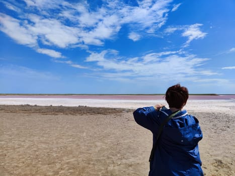 A individual stands on the sandy shore, photographing the vibrant pink salt flats beneath a bright blue sky.