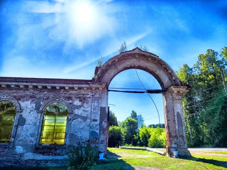 A crumbling brick archway with windows, surrounded by trees under bright, blue sky with the sun shining overhead