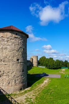 A medieval stone fortress with a tall round tower and extensive walls, set against a clear blue sky. The structure stands on a grassy area, evoking historical significance and rustic charm