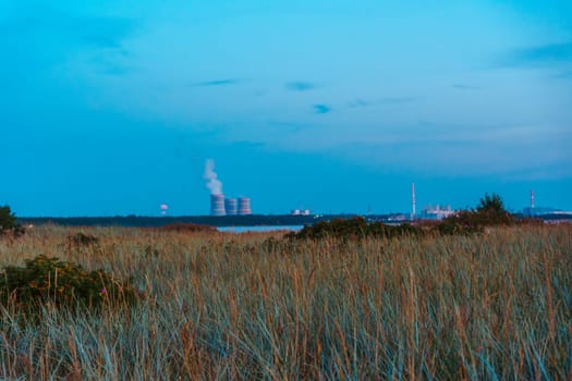 A distant industrial facility with smokestacks emitting smoke, seen across grassy dunes in the soft light of dusk. Calm and contrasting scene of nature and industry
